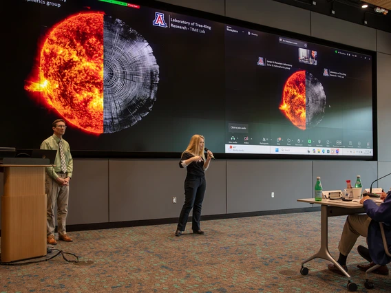 A woman speaks into a microphone while presenting in front of a large screen showing a composite image of the sun and a tree ring cross-section. A man stands beside a podium, and a seated judge listens from a table with microphones and water bottles. The presentation is part of the University of Arizona’s Big Ideas Challenge, hosted at the Grand Challenges Research Building.