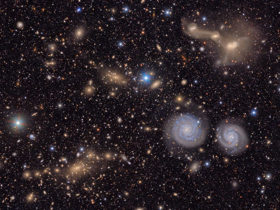 A sprawling, textured field of galaxies scattered across the deep black of space. It is filled with the delicate smudges and glowing cores of galaxies of many shapes, sizes and colors, as well as the bright multi-colored points of stars. To the lower left is a region filled with the hundreds of golden glittering gems of a distant galaxy cluster. In the foreground, below and right of center, two blue spiral galaxies look like eyes beneath the entangled mass of a triple galaxy merger in the upper right.