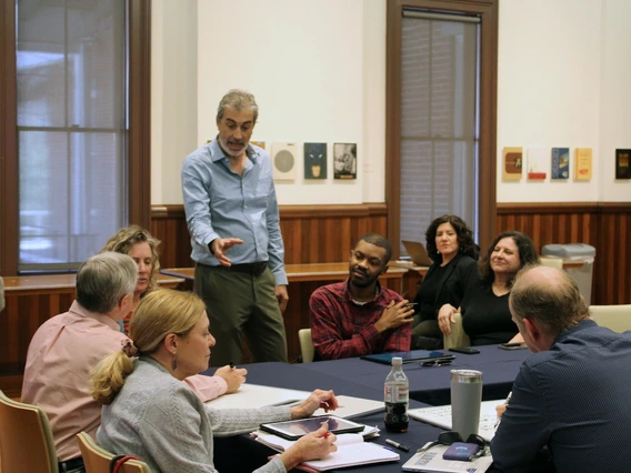 A man stands while speaking to a group at a round table during a strategic imperatives listening session. Participants are seated and appear engaged, with notebooks and tablets on the table.