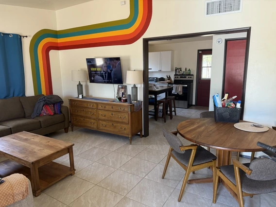 Living room with a brown couch, wooden coffee table, and a dresser with a mounted TV above it. A colorful retro stripe mural decorates the cream wall. A small round dining table with four chairs is also visible.