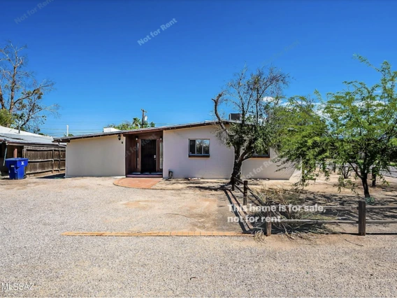A single-story, light-colored stucco house with a flat and slightly sloped roof, set against a clear blue sky. The front yard is mostly gravel with sparse desert vegetation, including a few small trees. A red brick path leads to a recessed front entry. There is a blue recycling bin on the left side near a wooden fence. The house has a minimalist desert-style appearance with no lawn.