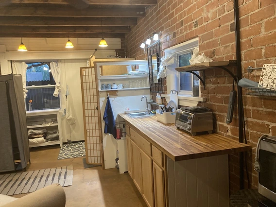Small kitchen space with wooden countertops, brick walls, and exposed ceiling beams. The area includes a sink, toaster oven, shelving with dishes, and pendant lighting overhead. A partition partially separates the kitchen from a windowed nook with shelving and curtains.