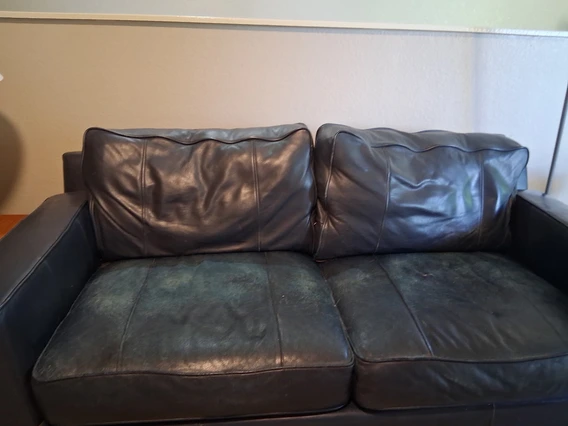 A worn black leather loveseat with visible discoloration and wear on the seat cushions. The back cushions appear in better condition, while the seating area shows fading and creases from use. The couch is placed against a beige wall with a white trim.