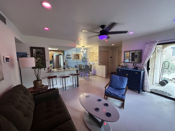 Modern living room with a green sofa, blue chair, and colorful ceiling lights, leading to a bright kitchen and outdoor area.