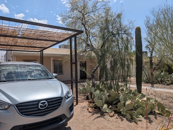 A single-story adobe-style house with a light tan exterior, shaded front patio, and desert landscaping including a large prickly pear cactus, a tall saguaro cactus, and several mesquite trees. A silver Mazda SUV is parked under a metal-frame carport with a slatted wooden cover. The sky is mostly clear with a few scattered clouds.