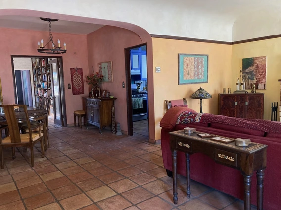 A cozy, Southwestern-style living and dining room with terra cotta tile floors, warm-toned stucco walls in shades of peach and yellow, and dark wood trim. The dining area features a round wooden table with chairs under a rustic chandelier. A hallway leads to a kitchen with blue cabinetry. The living area includes a red sofa, decorative pillows and blankets, and a Tiffany-style lamp on an end table. Framed art, plants, and vintage furniture pieces add charm to the space.