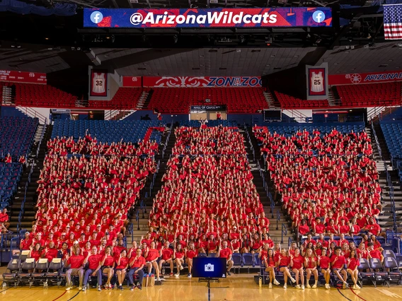 Hundreds of University of Arizona student-athletes wearing red shirts sit together in the shape of a large block “A” inside McKale Memorial Center during the Arizona Athletics welcome back BBQ. An American flag hangs above, and a scoreboard displays the @ArizonaWildcats handle.