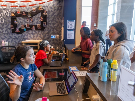 A pair of women wearing University of Arizona branded t-shirts sit the front desk of a student housing building. Standing in front of the desk are three female students wearing casual clothing.