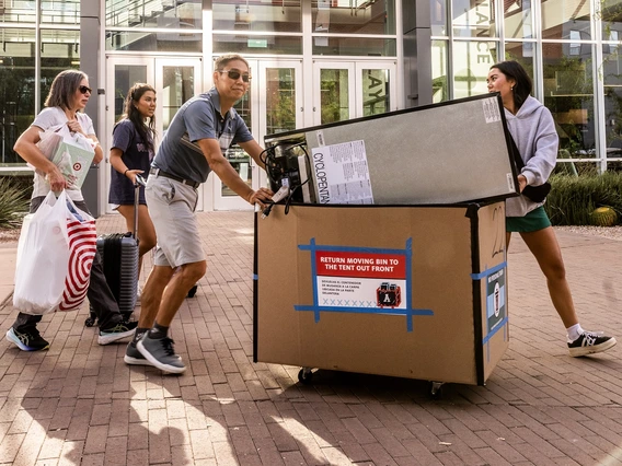 A middle aged man and woman help a pair of students push a large cardboard cart full of bedroom apparel and supplies into a college dorm. A large, glass-front building is in the background, with a large block "A" logo at the top.