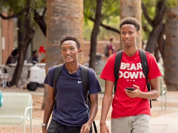 A man wearing black pants and a blue polo shirt and backpack walks next to a man wearing tan slacks and a red t-shirt that says "Bear Down." The two men are walking on a university campus with trees and seating in the background.