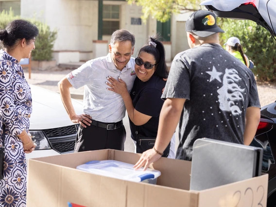 A man wearing a white, long sleeve button down greets a family outside of their vehicle. There is a large cardboard cart in front of the group containing a collection of dorm room decorations and supplies.