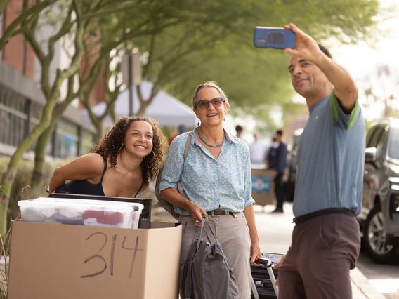 A young woman with curly brown hair poses for a selfie with an older woman wearing a light blue button down and grey slacks and a man wearing jeans and a blue t shirt. They are standing outside of a brick-built, college dorm building.