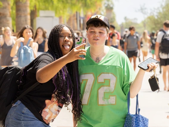A woman wearing denim jeans and a black t-shirt poses for a selfie next to a person wearing jeans and a light green jersey. They duo is standing on a college campus as other students walk by in the background.