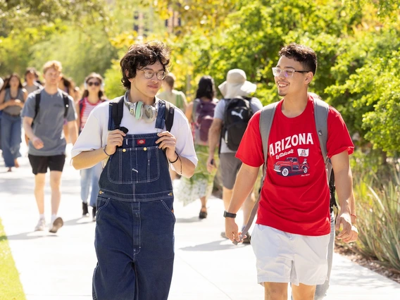 A man wearing overalls and a white t-shirt walks next to a man wearing white shorts and a red t-shirt, along a sidewalk on a university campus. There is grass, desert trees and other students in the background.
