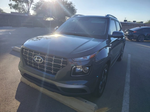 A dark gray Hyundai Venue parked in a lot, photographed from the front left. Sunlight creates a lens flare across the hood, highlighting the vehicle’s chrome-accented grille and sleek headlights. The background shows trees, a fence, and an empty parking area.