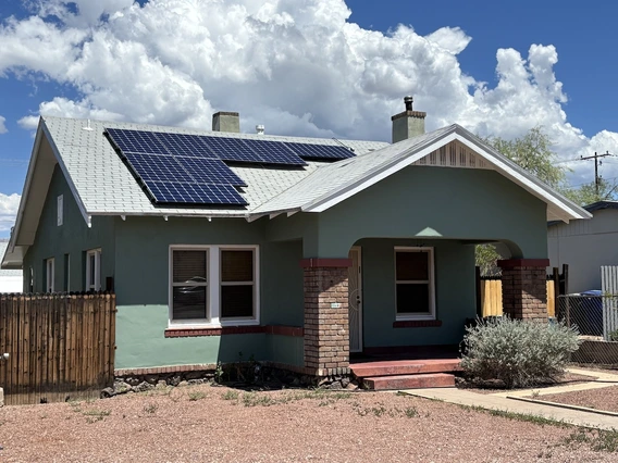 A green stucco bungalow with a white roof, red brick accents, and solar panels on the roof. The home has a covered front porch with brick columns and a gravel front yard under a partly cloudy sky.