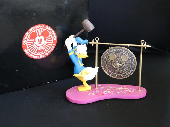 Figurine of Donald Duck wearing a blue sailor suit and hat, holding a large mallet above his head as if about to strike a hanging medallion. The medallion features Mickey Mouse’s face and reads “Official Disneyana Convention, Walt Disney World Resort, 1995.” The base of the figurine is pink with colorful confetti accents, and behind it is a black background with a red-and-white convention logo.