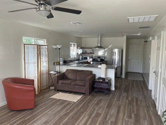 A cozy living space with a brown loveseat, red leather chair, and small tables. A ceiling fan is mounted above, and the kitchen is visible in the background with stainless steel appliances and granite counters.