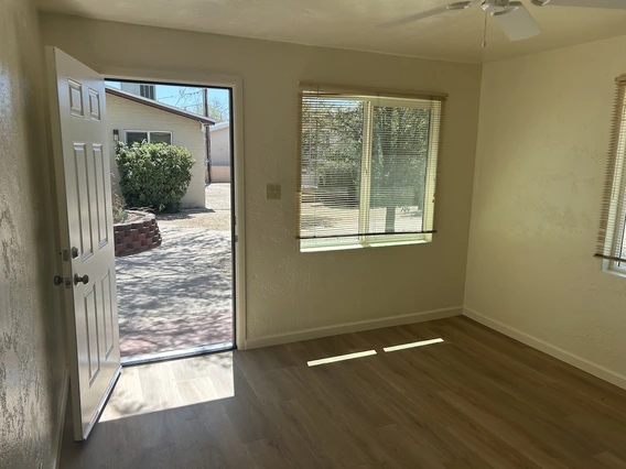 A bright room with light-colored walls and wood-style flooring. The front door is open, showing a small yard with a bush and a brick planter outside. Large windows let in natural light.