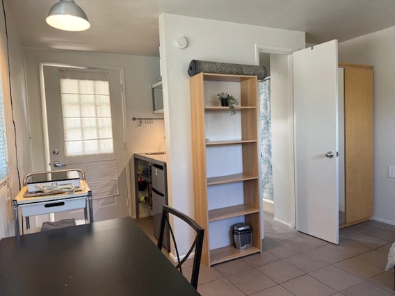 A small, tidy studio apartment with tile floors and white walls. The space features a black dining table with a chair, a metal utility cart with a microwave and hot plate, a tall wooden bookshelf with a plant and other items, and an open door revealing a bathroom with floral-patterned shower curtains. A yoga mat is rolled up on top of the bookshelf, and a wardrobe stands near the bathroom door. Natural light comes through a curtained door in the kitchenette area.