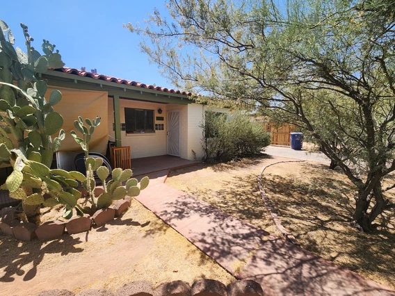 A small Southwestern-style house with a tiled roof, pale stucco walls, and a green-trimmed porch. A red walkway leads to the front door, which is shaded by a partial awning. The yard is landscaped with desert plants, including prickly pear cactus and mesquite trees, surrounded by dry soil and decorative stone borders. A wooden gate and a blue trash bin are visible in the background.