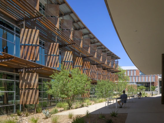 A modern architectural setting with two buildings facing each other. On the left, a multi-story structure showcases a series of vertical and horizontal rusty metal panels with glass windows underneath. The building on the right features a smooth, curved overhang extending above a walkway, offering shade. Below the left building, there is a landscaped area with a few small trees and desert plants. The sky is clear and bright blue.