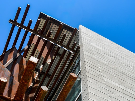 An upward view of a building corner against a vibrant blue sky. On the left, rusty metal beams project outward, forming a canopy-like structure. To the right, a concrete wall made of evenly stacked, uniform gray blocks extends upward, sharply intersecting with the metal and glass section. The composition highlights the geometric alignment and contrasting materials used in the building's architecture.