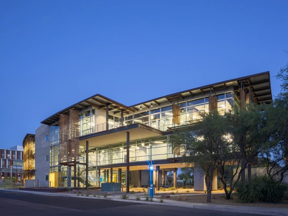 A modern building at dusk, featuring an extensive use of glass and steel. The structure is multi-leveled with large windows throughout, reflecting the clear blue sky. The building's exterior features a combination of exposed metal beams and panels. The brightly lit interior is visible through the glass, creating a warm contrast against the cool evening. In the foreground, there are landscaped areas with desert plants and a pathway leading to the entrance. 