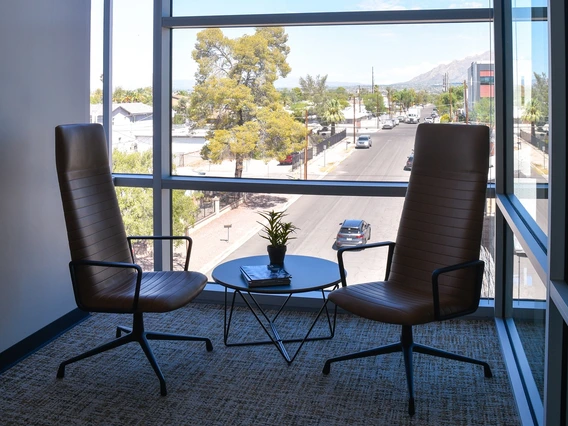 A modern office corner with floor-to-ceiling windows allowing a clear view of the street outside. Two brown leather chairs with metal frames are positioned on either side of a small round table. A small potted plant and a closed book or magazine rest on the table. Through the large windows, a sunny street scene is visible, featuring a wide road lined with trees and a few buildings. 