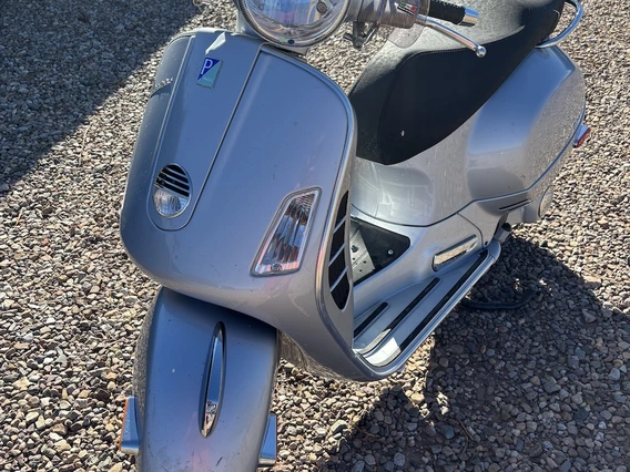 A silver Vespa scooter parked on a gravel driveway, shown from the front left angle. The headlight, handlebars, and front fender are visible, along with a small sticker near the headlight.