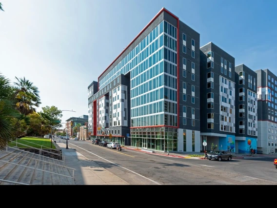 A modern multi-story apartment building with glass windows and dark gray paneling, located on a city street lined with palm trees.