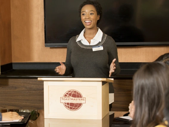 A woman stands at a podium, smiling and speaking to an audience.