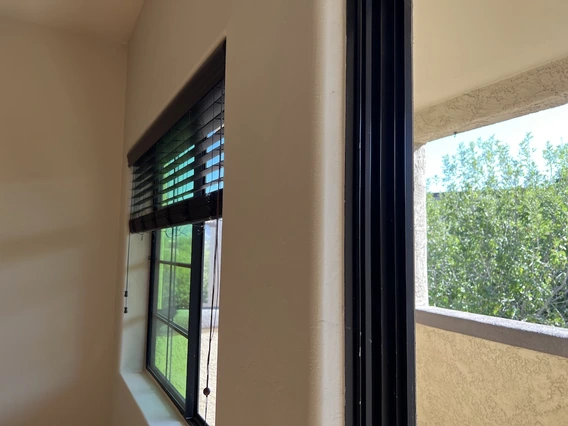 Interior view looking through a window with dark blinds partially raised, revealing sunlight, greenery, and part of the same stucco balcony outside.