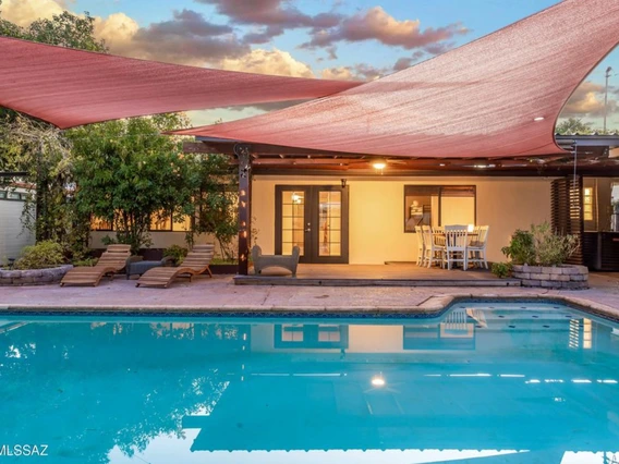 Backyard view of a home at sunset with a swimming pool in the foreground, several lounge chairs, and a covered patio area with string lights and dining furniture. Large triangular shade sails hang above the pool.