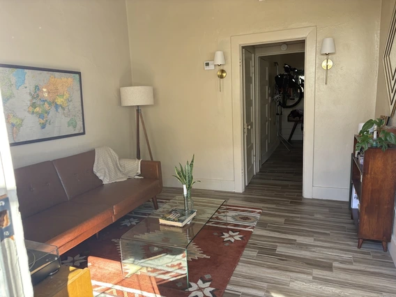 Cozy living room with a brown sofa, world map on the wall, and a floor lamp. A glass table on a patterned rug adds warmth, with plants and books.