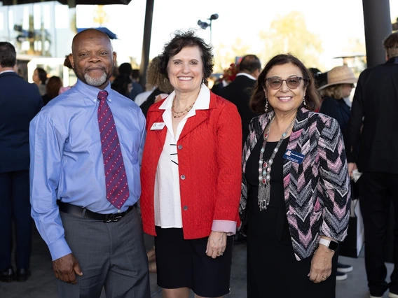 Three people stand together smiling at a reception outside the Stevie Eller Dance Theatre. The man on the left wears a blue shirt and red patterned tie, the woman in the center wears a red jacket, and the woman on the right wears a patterned jacket and glasses. Other guests mingle in the background.