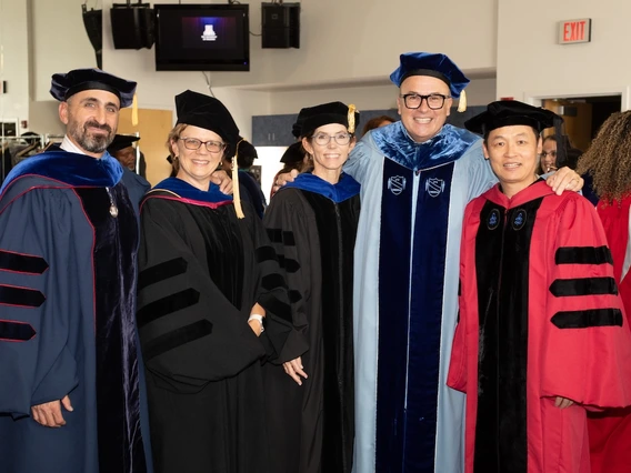 Five University of Arizona faculty members in academic regalia pose together and smile before a ceremony. They wear gowns in shades of blue, black, and red with velvet panels and caps, standing in a well-lit indoor space with other participants in the background.