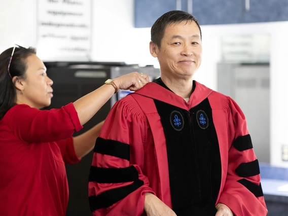 A woman helps a man adjust his academic regalia before a ceremony. The man, dressed in a red gown with black panels, smiles slightly while the woman, wearing a red top, focuses on the adjustment. The background shows an indoor space with soft lighting and blurred details.