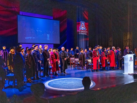 Faculty members in academic regalia stand on stage during the Luminaries Outstanding Faculty Awards ceremony at the University of Arizona. A large screen behind them shows a live feed of the event, and banners featuring the university’s logo hang on the stage.