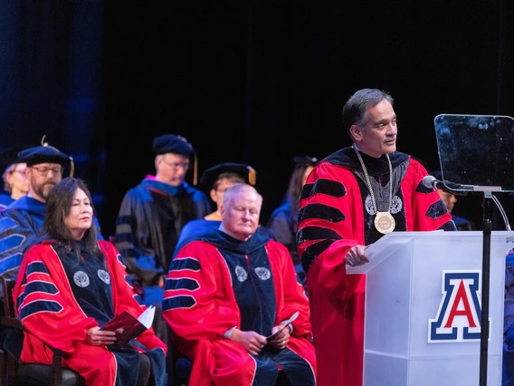 University of Arizona President Suresh Garimella speaks at a podium during the Luminaries Outstanding Faculty Awards ceremony. He wears academic regalia with a red gown and presidential medallion, with faculty members in similar attire seated behind him on stage.