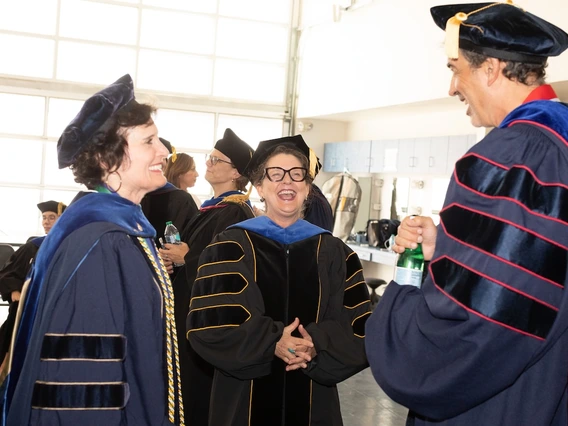 Three University of Arizona faculty members in academic regalia share a laugh before the Luminaries Outstanding Faculty Awards ceremony. Two hold water bottles as they chat in a brightly lit room with other participants in the background.