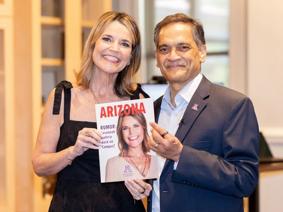 Two individuals are posing indoors. The woman on the left is wearing a black sleeveless dress and a necklace. She is smiling broadly while holding a magazine with a cover that features her photograph. The man on the right is wearing a dark suit with a light-colored shirt and is smiling while pointing at the magazine. The background consists of a softly focused interior with bookshelves and a window letting in natural light.