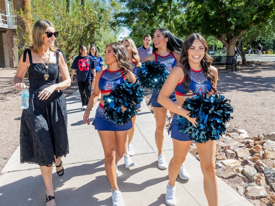 A group of cheerleaders walking along a pathway on a sunny day. There are four cheerleaders wearing matching blue and red uniforms with the letter 'A' on the front. They are holding shiny navy pom-poms and appear cheerful, engaging in conversation. To the left, a woman in a black dress with sunglasses holds a water bottle and smiles while talking to one of the cheerleaders. In the background, several other people are casually walking.