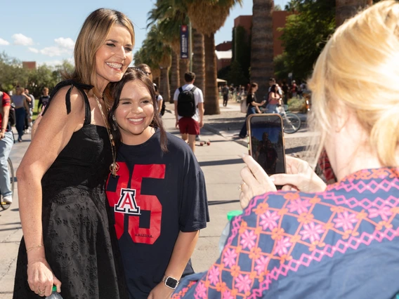 Two women are posing for a photograph on a sunny day. The woman on the left is wearing a black dress and has long hair. She is smiling broadly as she stands next to a young woman wearing a navy-blue T-shirt featuring a large red number "95" with the letter "A" and the word "ARIZONA" underneath. The young woman also smiles at the camera. A third person in the foreground, with blonde hair, is taking a photo with a smartphone.
