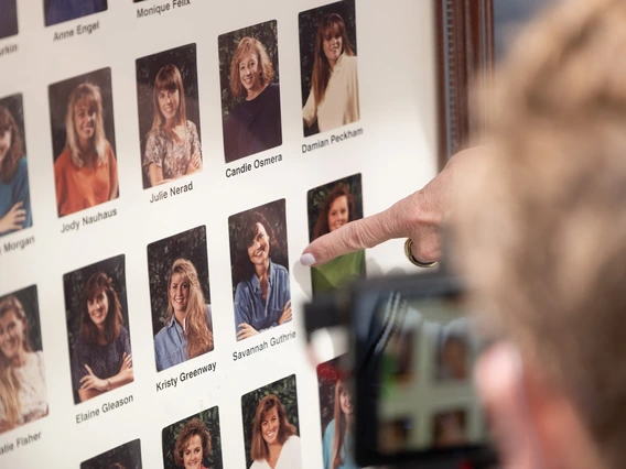 A collection of framed photographs featuring individual portraits arranged in a grid pattern. Each portrait is accompanied by a label with a name. The photographs depict people in various poses, mostly with smiles, set against a background of greenery. A person's finger, adorned with a ring, is pointing at one of the photographs in the middle of the frame.