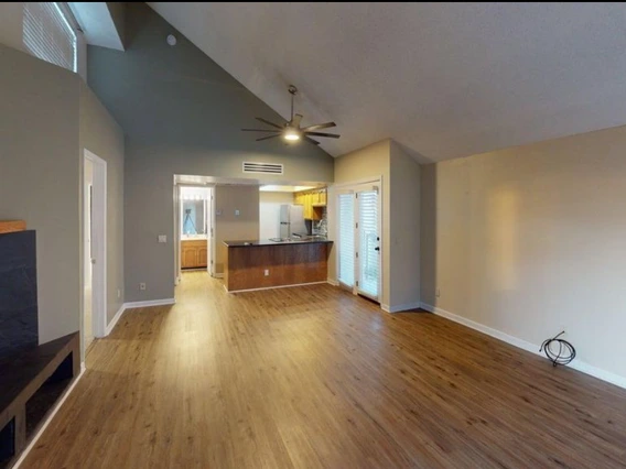 Open living area with wood-look flooring, high vaulted ceiling, and neutral walls. A ceiling fan hangs in the center, and French doors with blinds lead outside. The kitchen and hallway are visible in the background, along with a built-in wall mount for a TV on the left.