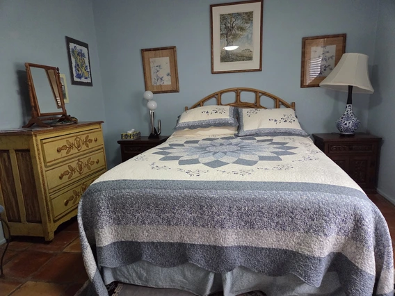 A cozy bedroom with a queen-size bed covered in a blue and white quilt, flanked by two dark wooden nightstands—one with a blue-and-white ceramic lamp and the other with a modern globe-style lamp. A yellow vintage dresser with decorative floral designs sits to the left, topped with a wooden mirror. Framed artwork hangs on the light blue walls above and around the bed.