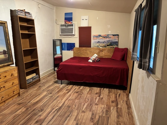 A simple bedroom with wood-look flooring, a twin bed covered in deep red bedding, and colorful artwork on the walls. A tall wooden bookshelf, a small dresser, and a floor mirror sit along one wall, while two windows with curtains bring in natural light.