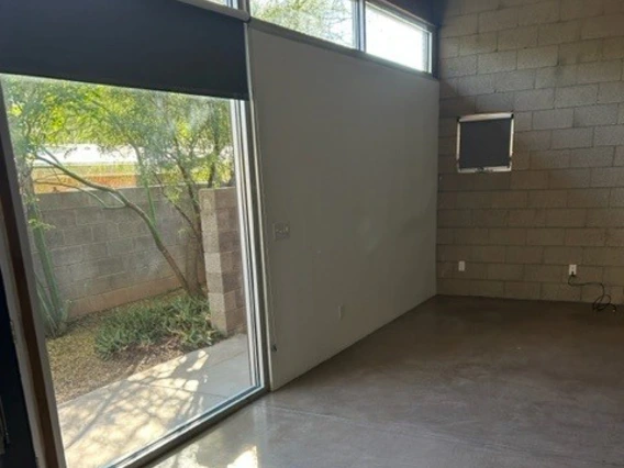 Interior of a modern room with polished concrete floors, tall narrow windows near the ceiling, and a large sliding glass door that opens to a small outdoor patio area with trees and a block wall.