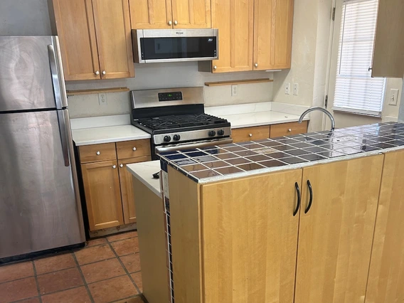 Kitchen with light wood cabinets, a stainless steel refrigerator and stove, a microwave mounted above the range, white countertops, and a small island with a dark tiled surface. The floor transitions from tile in the kitchen to carpet in the adjacent area.
